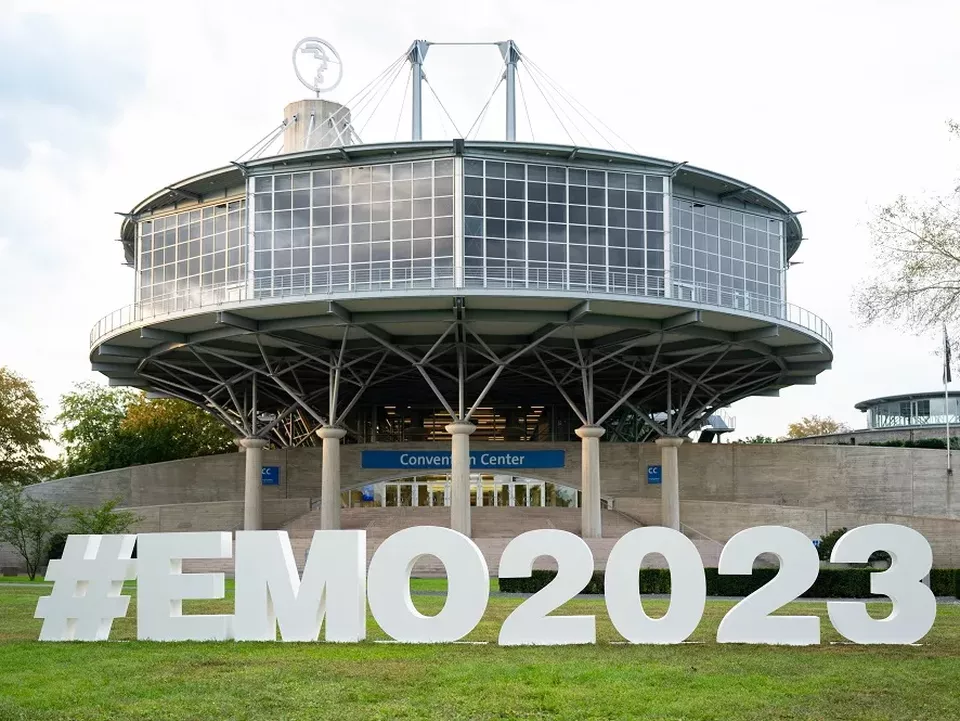 En rund byggnad på pelare med stora fönster som är entren till mässan med #EMO2023 som vita bokstäver på gräsmattan framför. 