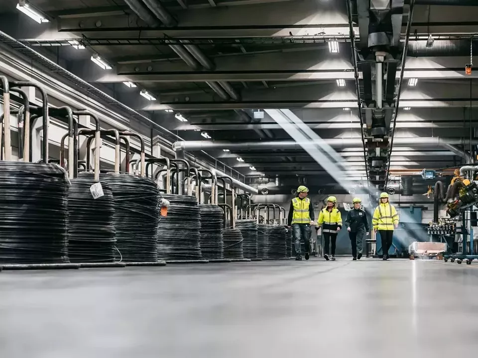 Fordonsunderleverantör tvingas varsla Fyra personer går i en industrilokal, tre i varseljacka, alla med skyddshjälm. På sidan av dem syns en lång rad med rullar av stål